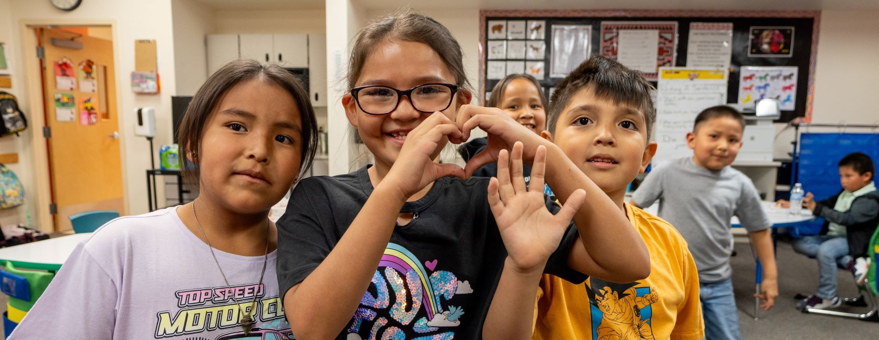 A group of smiling students in a classroom standing together. One student is making a heart with their hands.