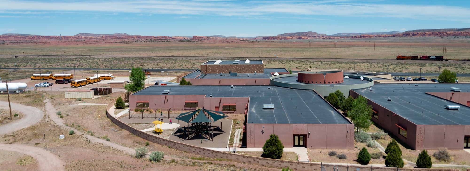 Aerial view of the school surrounded by a train and a vast desert landscape under a clear blue sky.